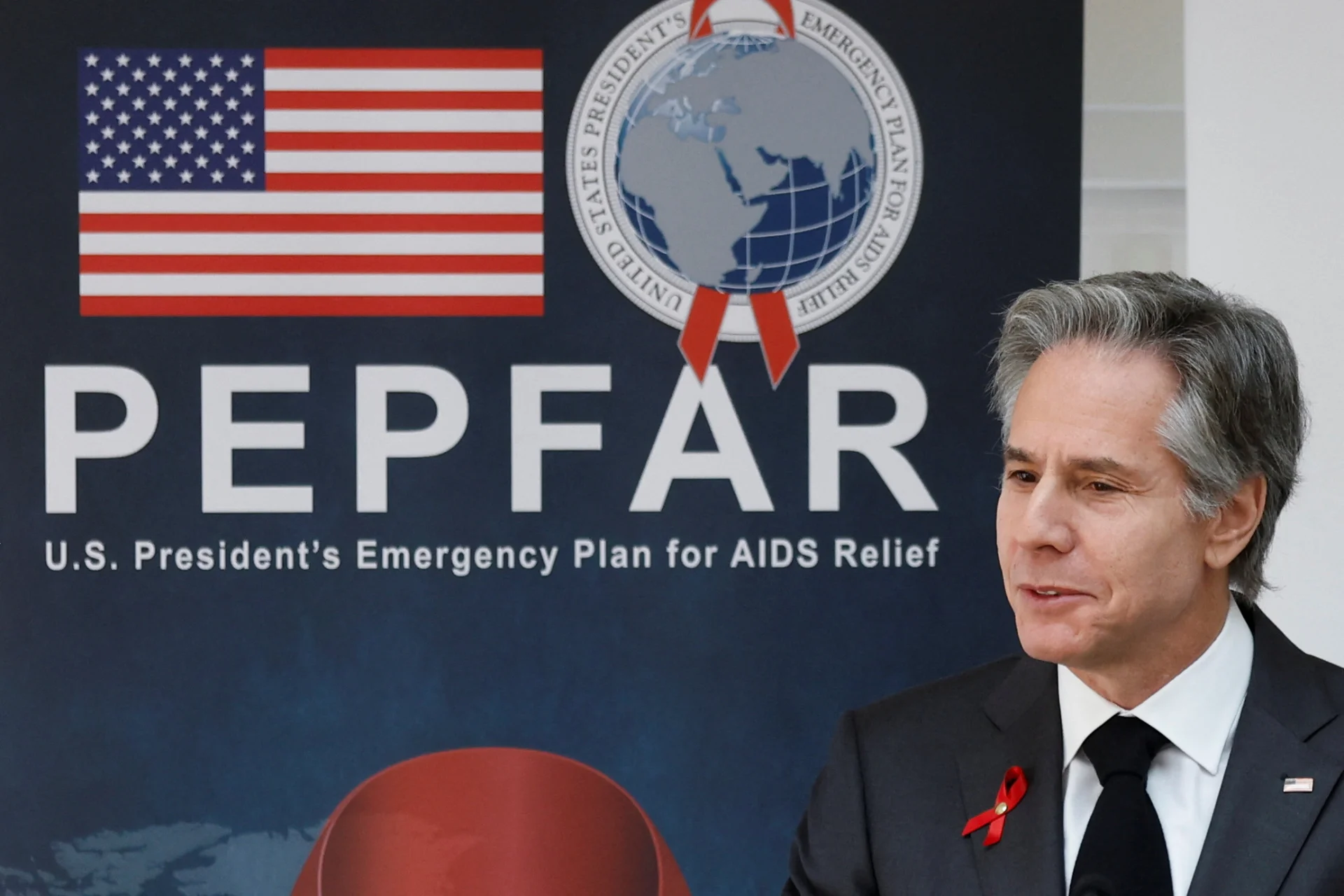 Man in a dark suit with a red ribbon stands beside a PEPFAR banner featuring the US flag and a globe emblem.