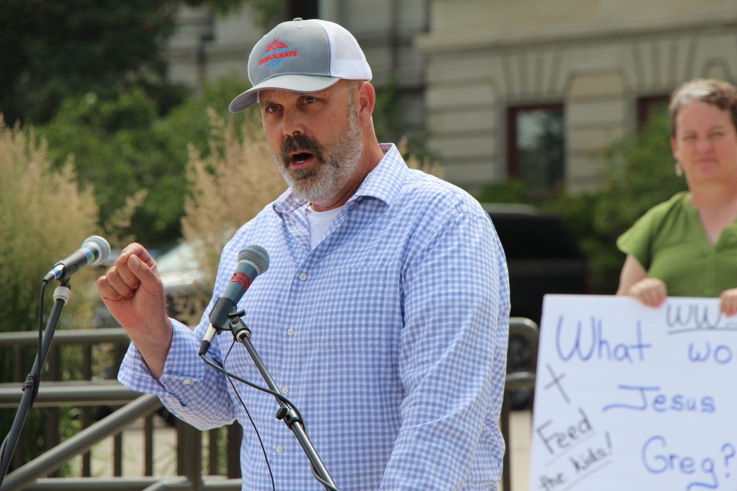 Man speaking at an outdoor event into multiple microphones, wearing a gray cap and blue checkered shirt.