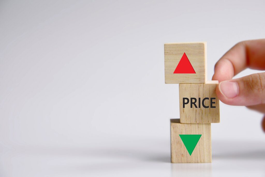 A hand stacks three wooden blocks displaying a red upward arrow, the word "PRICE," and a green downward arrow, representing fluctuating prices.