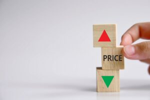 A hand stacks three wooden blocks displaying a red upward arrow, the word "PRICE," and a green downward arrow, representing fluctuating prices.
